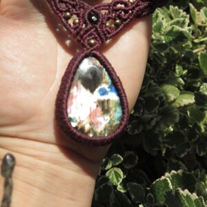 May include: A maroon macrame necklace with a large iridescent gemstone pendant. The pendant is teardrop shaped and has a variety of colors, including blue, green, and pink.