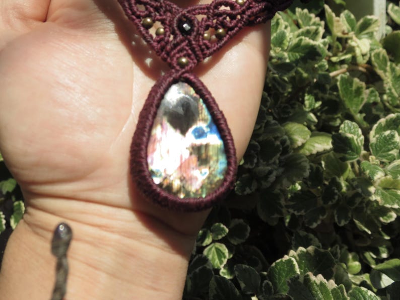 May include: A maroon macrame necklace with a large iridescent gemstone pendant. The pendant is teardrop shaped and has a variety of colors, including blue, green, and pink.