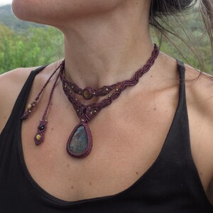 May include: A maroon macrame choker necklace with a teardrop-shaped blue gemstone pendant. The necklace has two strands and features small gold beads.