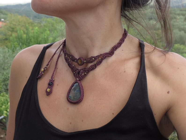 May include: A maroon macrame choker necklace with a teardrop-shaped blue gemstone pendant. The necklace has two strands and features small gold beads.