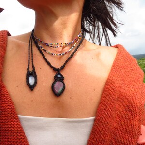 Puede incluir: Una colección de collares hechos a mano con intrincados diseños de macramé. Los collares incluyen hilos de cuentas de colores y colgantes con piedras iridiscentes, todo sobre un cordón negro. La persona lleva una rebeca naranja.