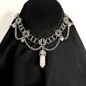 Puede incluir: Un collar plateado con un diseño en capas. El collar presenta una serie de eslabones de cadena interconectados y cadenas colgantes. Pequeñas gemas rosas están intercaladas en el diseño, y un colgante de cristal rosa cuelga del centro.