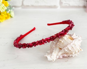 May include: A thin red headband adorned with small, irregular, deep red stone chips along the top surface. The headband is resting on a white, textured seashell, showcasing the vibrant red colour and the textured finish of the stone embellishments.