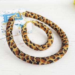 May include: A beaded necklace and bracelet set with a leopard print pattern in shades of brown, black, and gold. The jewelry is displayed on a white surface, with a small gift box in the background.
