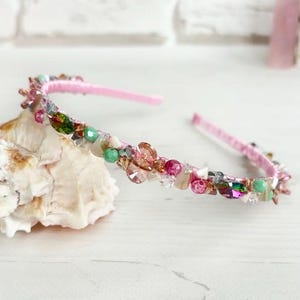 May include: A pink headband adorned with an array of colorful beads and crystals. The decorative embellishments include shades of pink, green, and clear crystals. The headband is resting on a white seashell.