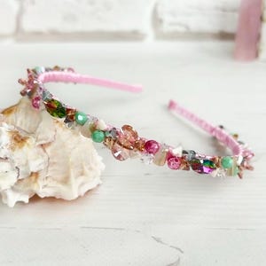 May include: A pink headband adorned with an array of colorful beads and crystals. The decorative embellishments include shades of pink, green, and clear crystals. The headband is resting on a white seashell.
