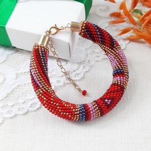 Könnte beinhalten: Ein Armband aus roten, goldenen, lila und blauen Perlen. Das Armband besteht aus kleinen, runden Perlen und hat einen goldfarbenen Verschluss. Das Design zeigt ein Spiralmuster mit einer Mischung aus Farben.