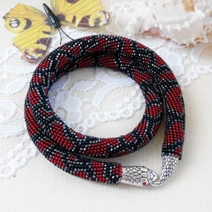 Peut inclure: Un collier de perles en forme de serpent. Le collier présente un motif de perles rouges, noires et argentées. Le fermoir est une tête de serpent argentée avec des yeux rouges. Le collier est présenté sur une surface en dentelle blanche.