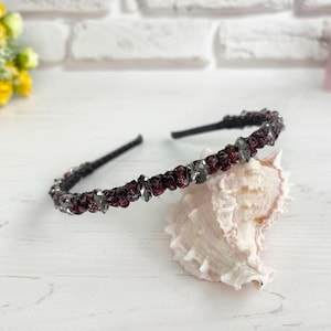 May include: A black headband adorned with burgundy and gray crystals. The decorative hair accessory is set against a white background with a seashell. The headband is a stylish hair accessory.