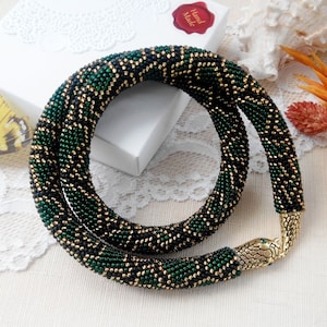 Puede incluir: Un collar de cuentas con cierre de serpiente. El collar presenta un patrón de cuentas verdes, negras y doradas. El cierre es de metal dorado con forma de cabeza de serpiente y ojos verdes. El collar se exhibe sobre una superficie blanca.