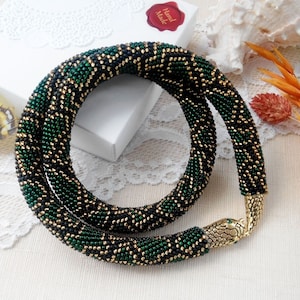 Puede incluir: Un collar de cuentas flexible con diseño tubular y un patrón repetitivo de cuentas de semillas negras, verde oscuro y oro metálico. El collar termina en un cierre de metal dorado con forma de cabeza de serpiente y pequeños ojos verdes.