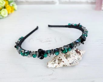 May include: A black headband embellished with a series of faceted beads in shades of emerald green, dark grey, and black. The beads are arranged in a continuous row along the top of the thin, flexible band, creating a textured, sparkling appearance.