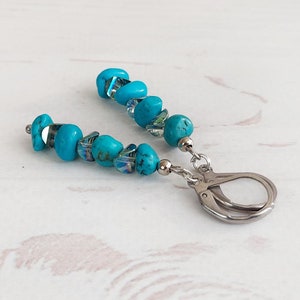 May include: Turquoise beaded earrings with a silver clasp. The earrings feature small, irregular turquoise beads interspersed with clear, iridescent beads. The silver clasp is a simple, secure design. The earrings are displayed on a white wooden surface.