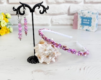May include: A set consisting of a headband and matching dangle earrings, both featuring small, irregular pink and purple beads. The earrings hang from a black metal display stand, while the headband rests on a white seashell.