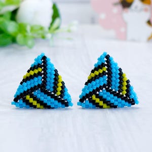 May include: A pair of handmade triangle-shaped earrings. The earrings are made of small, colorful beads in a geometric pattern of blue, black, and yellow. The earrings are displayed on a white surface, with flowers and a gift box in the background.