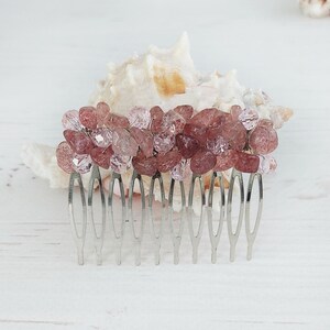 Könnte beinhalten: Ein Haarkamm mit einem dekorativen Oberteil, das mit Clustern aus kleinen, facettierten, durchscheinenden rosa und dunkelrosafarbenen Perlen verziert ist. Der Kamm selbst besteht aus silberfarbenem Metall mit mehreren dünnen, gebogenen Zinken.