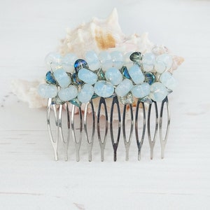 May include: A hair comb featuring a decorative top section adorned with clusters of light blue, translucent, and dark blue beads. The comb has a silver-toned metal base with multiple prongs designed for securing hair, showcasing a beaded arrangement.