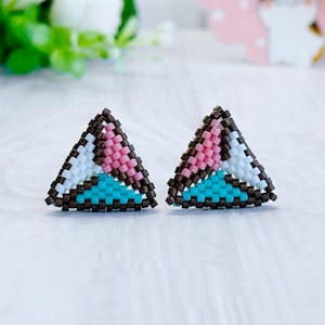 Könnte beinhalten: Ein Paar dreieckige Ohrstecker aus kleinen Perlen. Jeder Ohrstecker zeigt ein geometrisches Muster, das in drei Abschnitte unterteilt ist, die in Pink, Weiß und Türkis gefärbt sind und mit dunkelbraunen Perlen umrandet sind.
