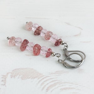May include: A pair of dangle earrings featuring a combination of pink and red-toned gemstone beads. The beads are strung together and attached to silver-colored leverback earring hooks. The earrings are displayed on a white wooden surface.