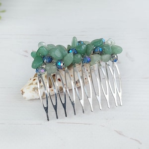 Op de afbeelding: Een haarspeld met zilveren metalen tanden, versierd met groene en blauwe edelstenen. De edelstenen zijn gegroepeerd en vormen een decoratief accent. De kam rust op een zeeschelp, wat een strand- of oceaanthema suggereert.