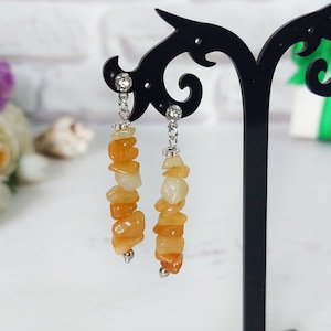 Puede incluir: Un par de pendientes colgantes con fragmentos de piedra irregulares en tonos naranja y crema. Las piedras están ensartadas en un alambre de metal plateado con pequeños cristales transparentes en la parte superior y una cuenta plateada al final.