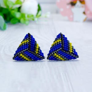 May include: A pair of triangular stud earrings crafted from small beads in a geometric pattern. The design features alternating diagonal stripes of vibrant blue, bright yellow, and metallic dark gray beads, creating a textured, three-dimensional look.