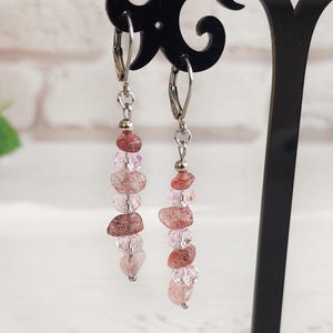 Peut inclure: Boucles d'oreilles pendantes avec fermoir à levier. Les boucles d'oreilles présentent une combinaison de pierres précieuses roses et brun rougeâtre et de perles transparentes. Les boucles d'oreilles sont exposées sur un présentoir noir, avec un arrangement floral flou en arrière-plan.