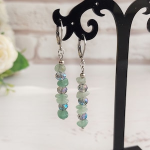 Może przedstawiać: Para kolczyków wiszących z haczykami w kolorze srebrnym. Każdy kolczyk ma pionowy układ jasnozielonych, nieregularnych koralików i iryzujących, okrągłych koralików. Kolczyki są prezentowane na czarnym stojaku na rozmytym tle.