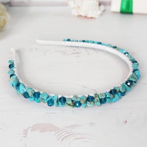May include: A white headband adorned with turquoise and teal-colored stones. The stones are irregularly shaped and clustered along the band, creating a textured and decorative accessory. The headband is set against a light-colored background.