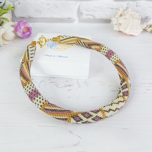 Puede incluir: Un collar de cuentas con un cierre dorado. El collar presenta un diseño en espiral con secciones alternas de cuentas blancas, doradas, negras y rosas. El collar se exhibe sobre una superficie blanca con flores en el fondo.