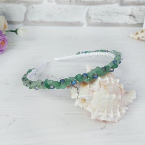 May include: A headband featuring a white base adorned with small, irregular green stone chips and interspersed dark blue faceted beads. The accessory is curved and displays a textured, multi-faceted surface created by the arrangement of the stones.