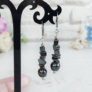 May include: A pair of drop earrings featuring dark gray, metallic-looking hematite beads in various shapes, including small cubes and a larger round bead at the bottom. The beads are strung on silver-toned metal wire with lever-back ear wires.