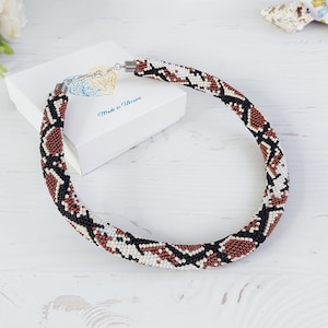 Op de afbeelding: Een kralenketting met een slangenpatroon in bruin, zwart en wit. De ketting is cirkelvormig en ligt op een wit houten oppervlak. Een witte doos en bloemen zijn op de achtergrond. De tekst "Made in Ukraine" is zichtbaar.