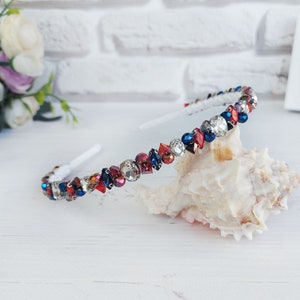 Puede incluir: Una diadema blanca adornada con pedrería roja, azul y transparente. Las piedras decorativas están engarzadas en una banda plateada. La diadema se exhibe sobre una concha, con un fondo de ladrillos blancos.