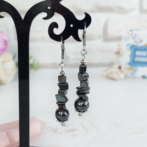 May include: A pair of dangle earrings featuring dark gray, geometric-shaped beads and a large, round bead at the bottom. The earrings are suspended from silver-colored ear wires. The earrings are displayed on a black stand against a soft background.