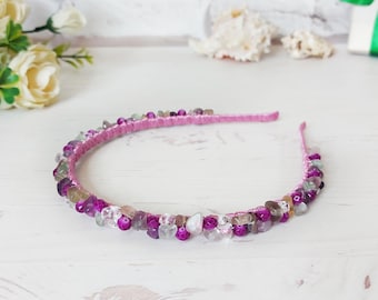May include: A pink headband embellished with a variety of small, polished, multi-coloured stones in shades of purple, magenta, and clear white. The stones are securely attached along the entire length of the thin, flexible pink band.