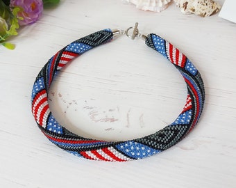 Può includere: Una collana di perline con un motivo ripetuto di strisce e stelle rosse, bianche e blu, che ricorda la bandiera americana. Il design tubolare è realizzato con piccole perline di vetro e fissato con una chiusura a T color argento.