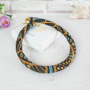 May include: A tubular beaded necklace featuring a geometric pattern in black, gold, and turquoise seed beads. The necklace is constructed with a flexible, rounded design and includes a gold-toned metal toggle clasp closure at the ends.