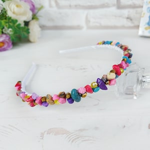 May include: A white headband adorned with colorful, irregularly shaped stones in shades of pink, red, blue, purple, and gold. The stones are arranged around the band, creating a decorative accessory.