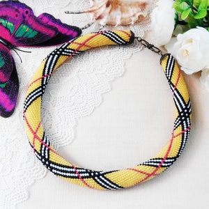 May include: A circular beaded necklace featuring a yellow, black, white, and red plaid pattern. The necklace is constructed from small, uniform beads arranged in a tubular shape, secured with a dark metal clasp at the back of the neck.