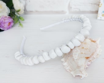 May include: A white headband featuring a continuous row of smooth, rounded white beads. The headband is positioned resting against a textured, light-coloured seashell, with a small floral arrangement visible in the upper left corner.