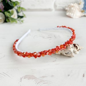 May include: A white headband embellished with a dense arrangement of small, irregular orange and coral-colored stone chips interspersed with translucent, faceted orange beads that catch the light along the entire length of the band.
