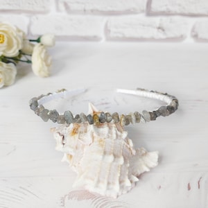 May include: A white headband adorned with a continuous row of small, irregular, grey and translucent stone chips. The headband is positioned resting on a white, textured seashell, showcasing the natural variation in the stone colors and shapes.
