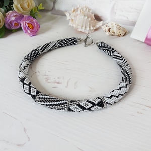 Puede incluir: Un collar tubular de cuentas con un patrón geométrico repetitivo en negro, blanco y plata. El collar está construido con un diseño flexible y redondeado, y se asegura con un cierre de palanca de metal en tono plateado.