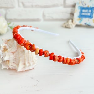 May include: A white headband adorned with vibrant orange coral beads. The coral is strung along the band, creating a natural and eye-catching accessory. The headband is a stylish hair accessory.