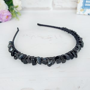 May include: A black headband adorned with a series of polished, irregular black stone chips secured along the band. The stones have a glossy, reflective surface and are arranged in a continuous row, creating a textured appearance across the accessory.