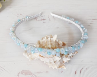 May include: A headband wrapped in white fabric and adorned with a series of small, polished light blue and translucent white stone chips arranged along the entire outer curve of the band, creating a textured and multi-tonal appearance.