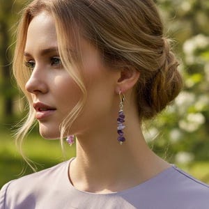 May include: A pair of drop earrings featuring a vertical arrangement of small, irregular, polished purple amethyst stone chips strung on a thin silver-toned metal wire, worn with a light lavender long-sleeved top with a round neckline.