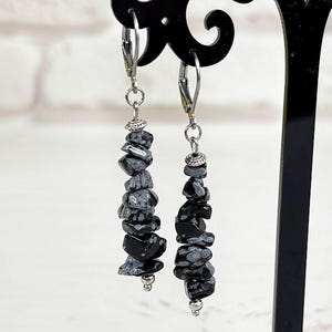 May include: Black and gray stone earrings hanging from a black display stand. The earrings are made of small, irregularly shaped stones strung together. The stand is on a white surface, with a bouquet of yellow flowers and a sign in the background.