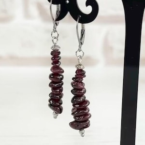 May include: A pair of dangle earrings with dark red, stacked stone beads. The earrings are displayed on a black, decorative stand. The background includes a bouquet of yellow and white flowers and a sign that says "Made in Ukraine."